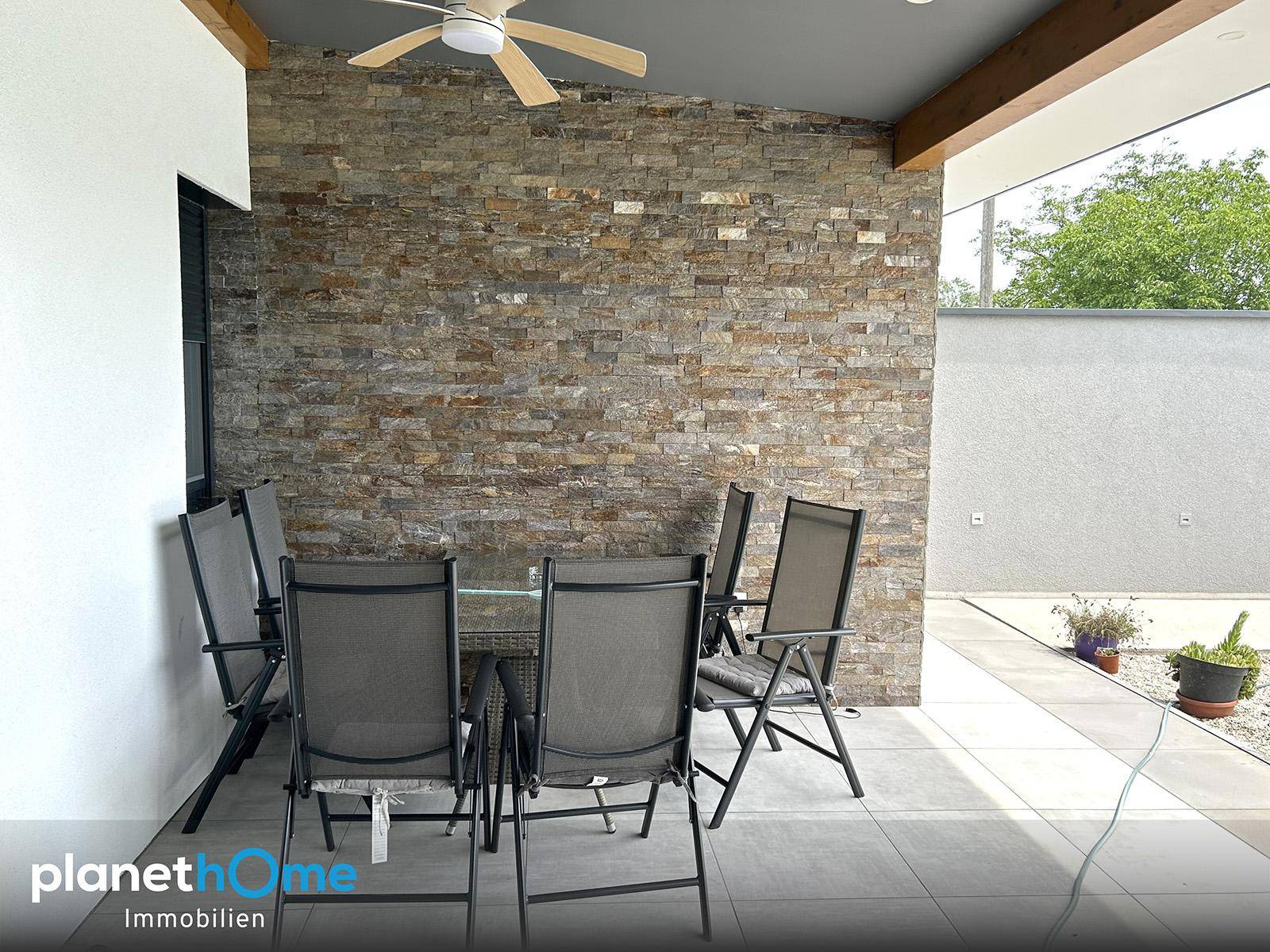 Überdachte Terrasse mit Sitzgelegenheiten, Deckenventilator und dekorativer Steinwand, ideal zum Entspannen.