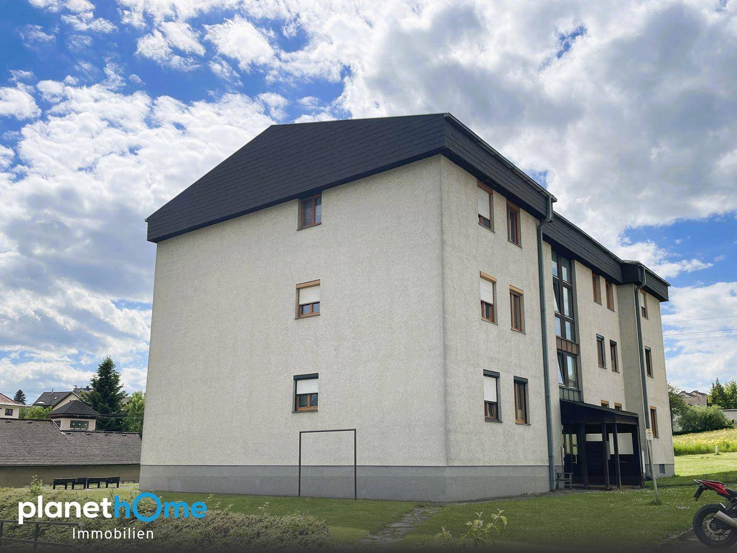 Mehrstöckiges Wohngebäude mit heller Fassade und dunklem Dach, umgeben von Grünflächen.