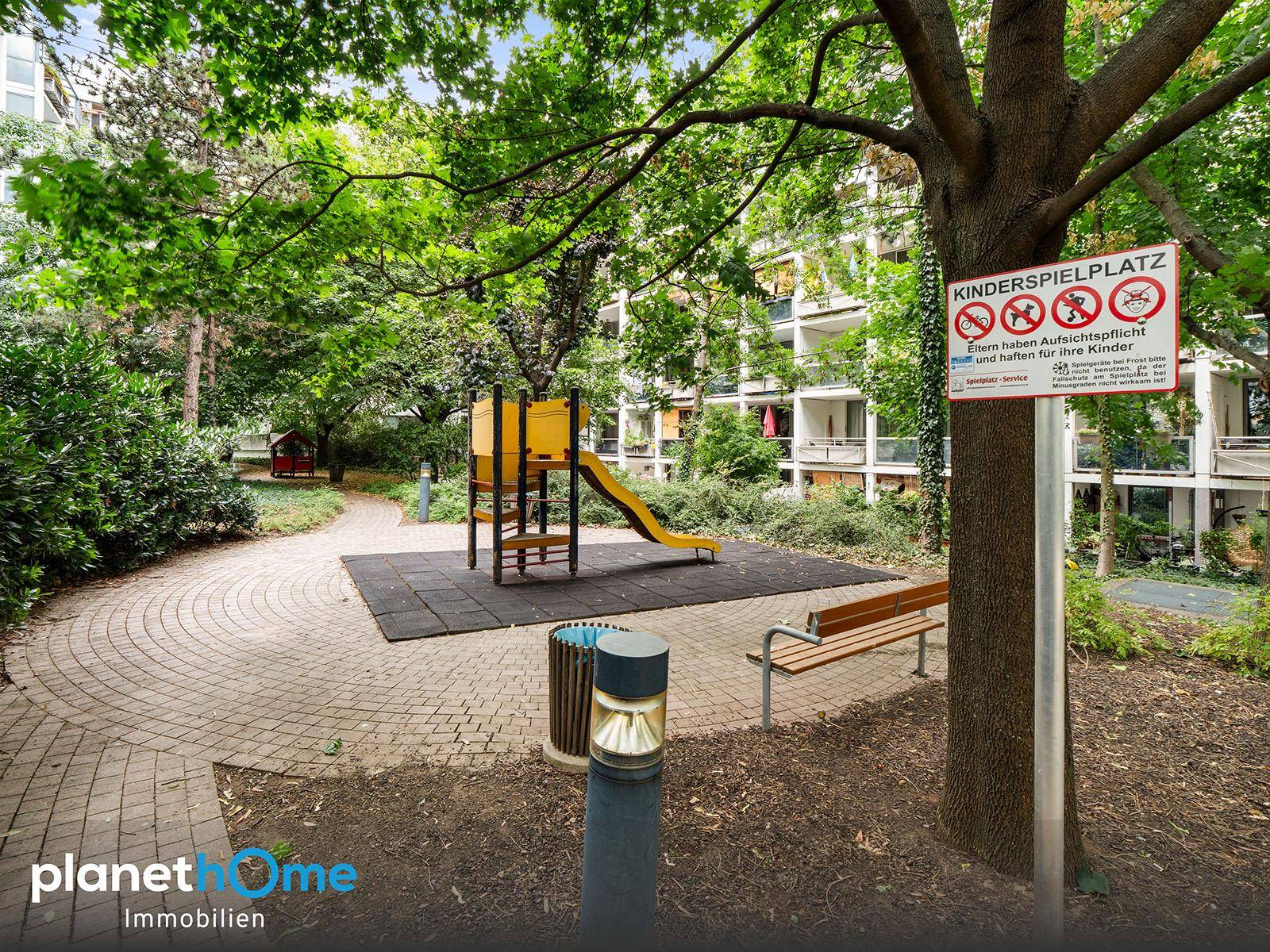 Grüner Kinderspielplatz mit Rutsche und Sitzgelegenheiten, umgeben von Bäumen.