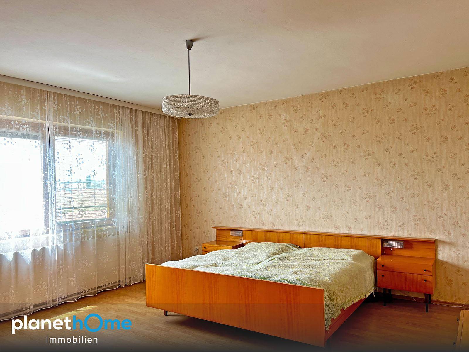 Schlafzimmer mit Doppelbett, gemusterter Tapete und großem Fenster, das viel Tageslicht hereinlässt.