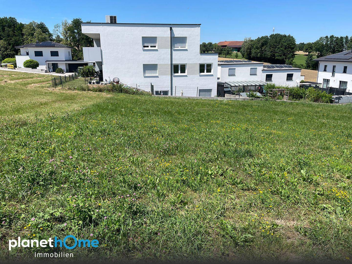 Blick über ein grünes Baugrundstück auf ein modernes Mehrfamilienhaus und weitere Wohnhäuser.