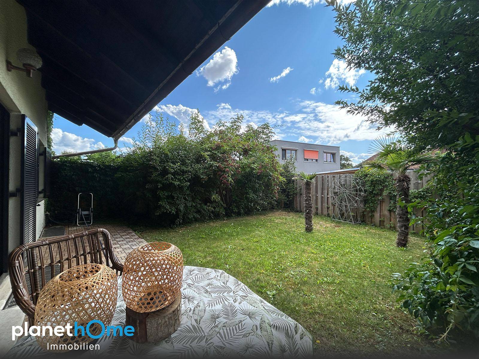 Blick vom überdachten Terrassenbereich in den Garten mit Rasen, Hecken und Palmen.