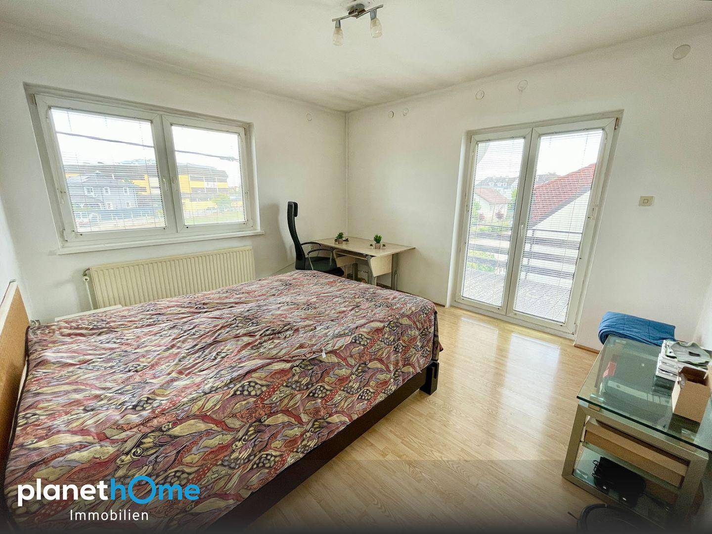 Schlafzimmer mit Zugang zum Balkon, Laminatboden und einem Schreibtisch am Fenster.