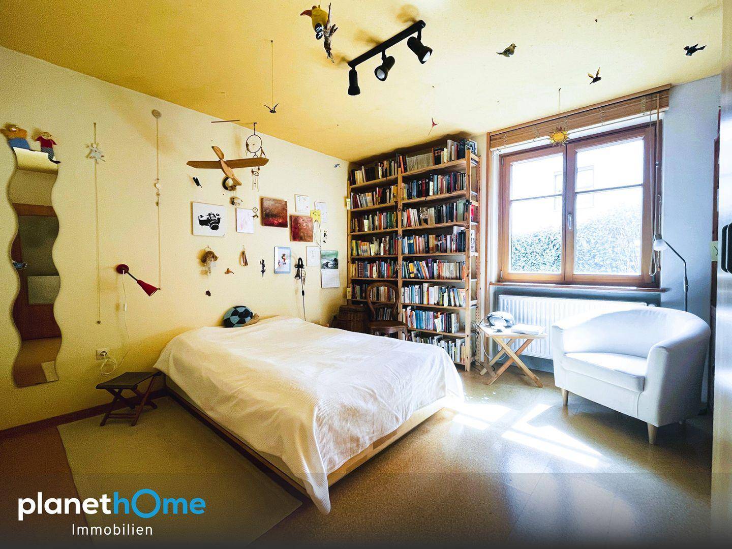 Helles Schlafzimmer mit Einzelbett, großem Bücherregal und Blick ins Grüne durch das Fenster.