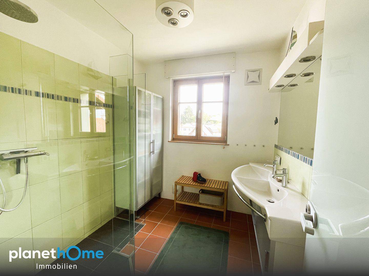 Badezimmer mit grünen Wandfliesen, Dusche, Fenster und einem Doppelwaschbecken mit Spiegel.