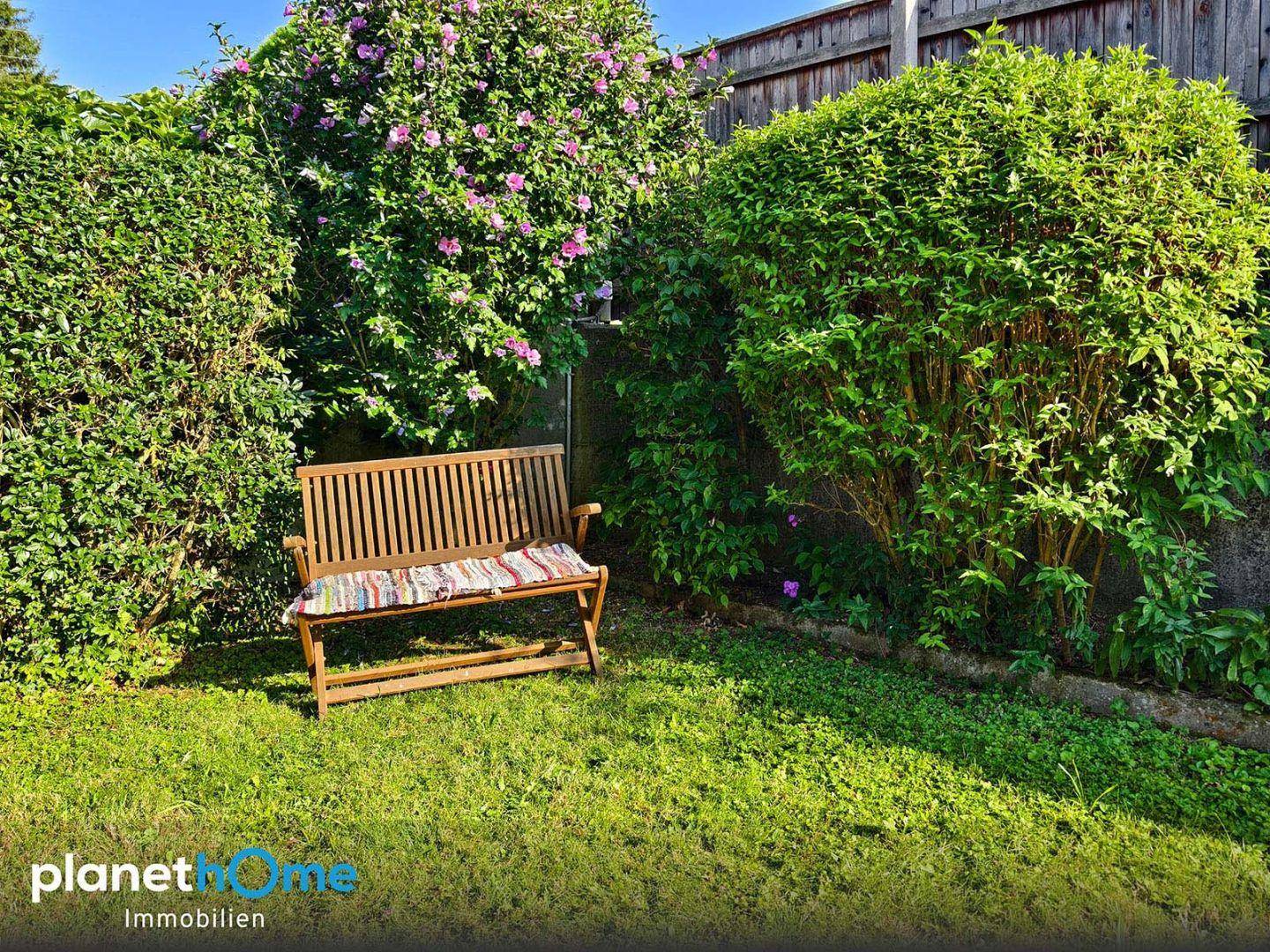 Ruhige Sitzecke im Garten mit Holzbank, umgeben von dichten Hecken und blühenden Sträuchern.
