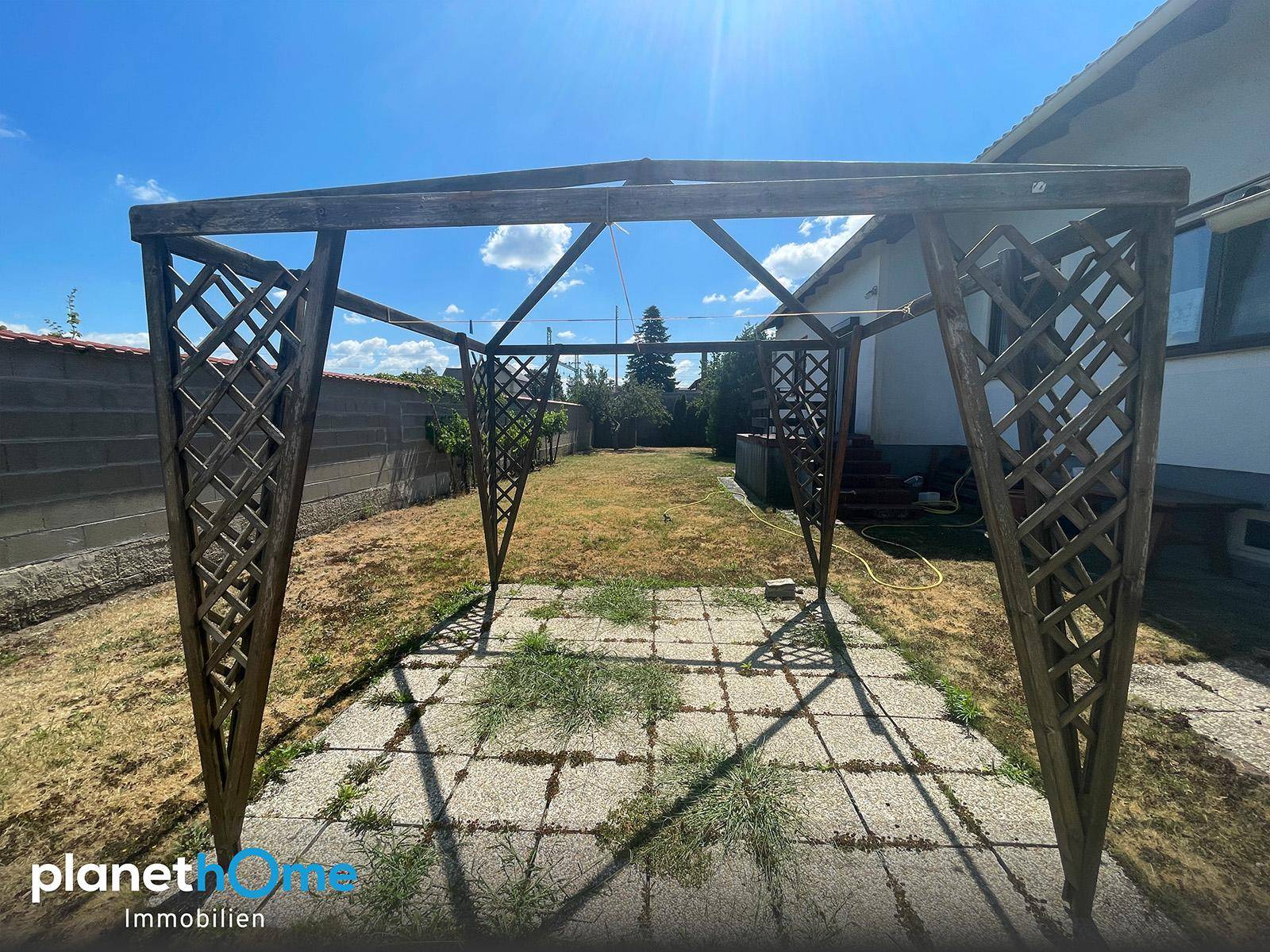 Gartenbereich mit einer Holzpergola und gepflastertem Boden unter blauem Himmel.