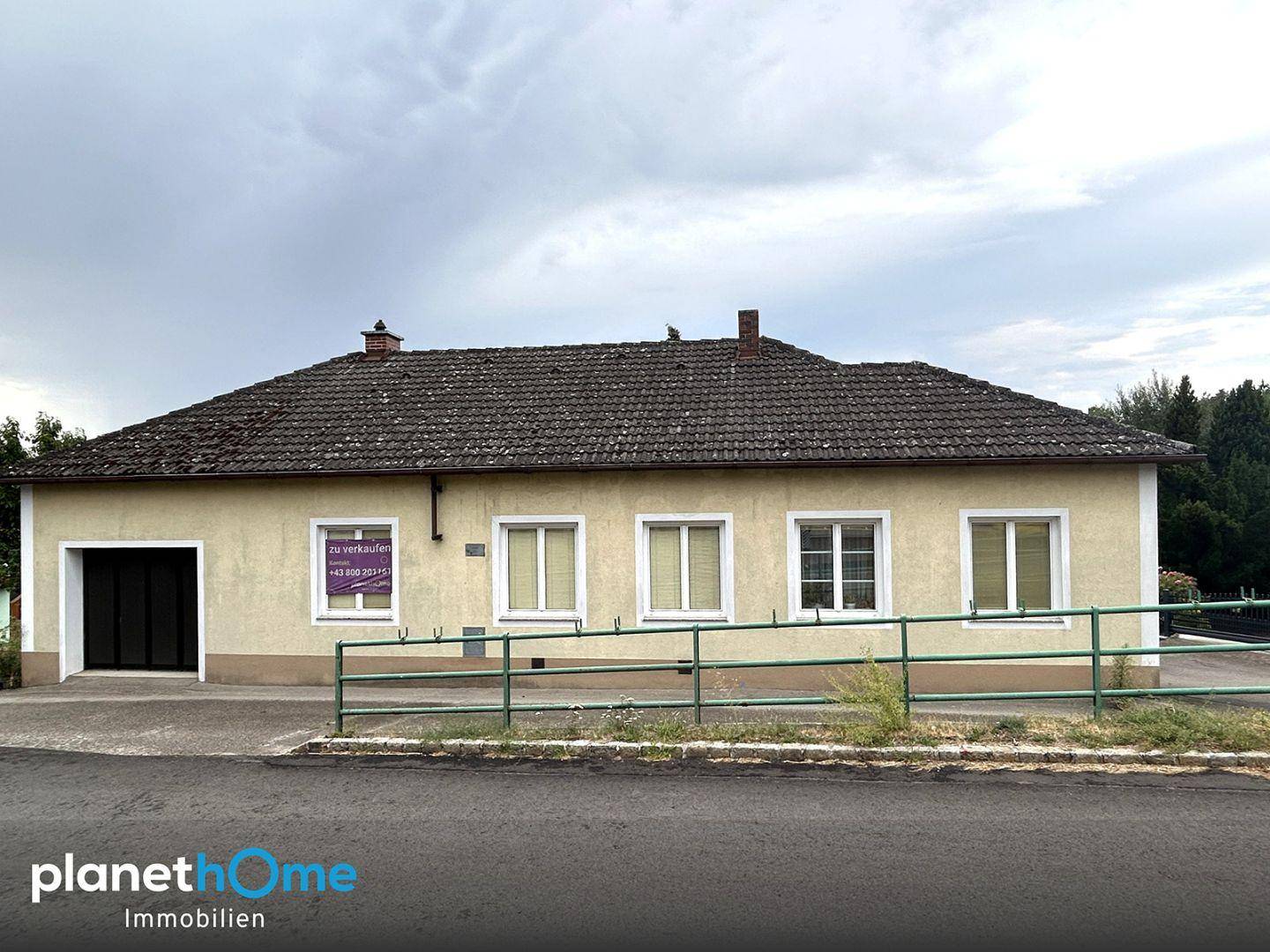 Einfamilienhaus mit heller Fassade, Satteldach und Garage, umgeben von einem grünen Zaun.
