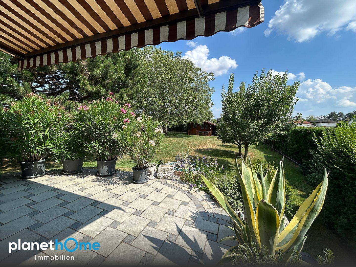 Gepflasterte Terrasse mit Markise, Pflanzen und weitem Blick in den Garten.