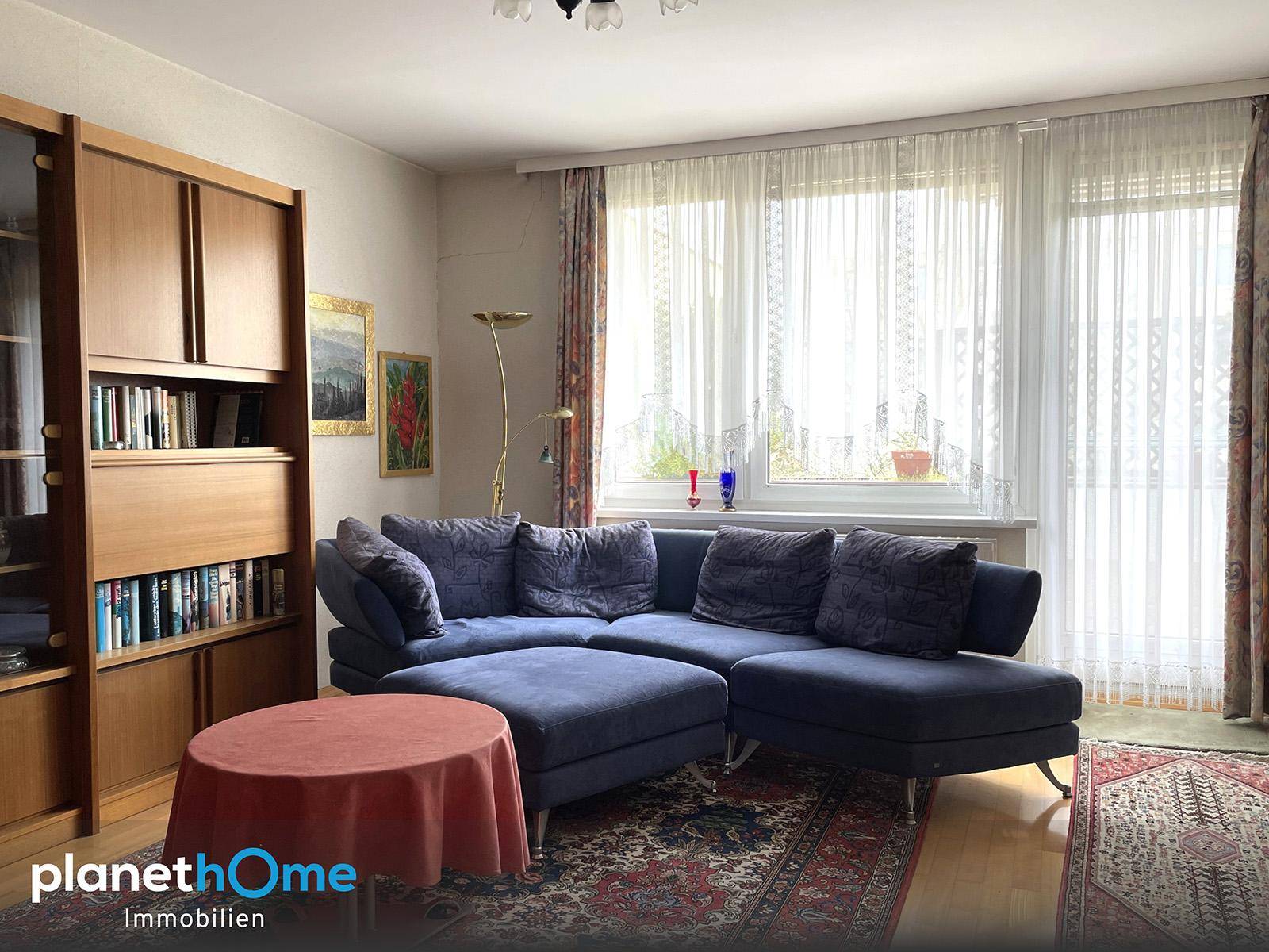 Gemütliches Wohnzimmer mit großem blauen Sofa, Holzregal und hellen Fenstern mit Vorhängen.