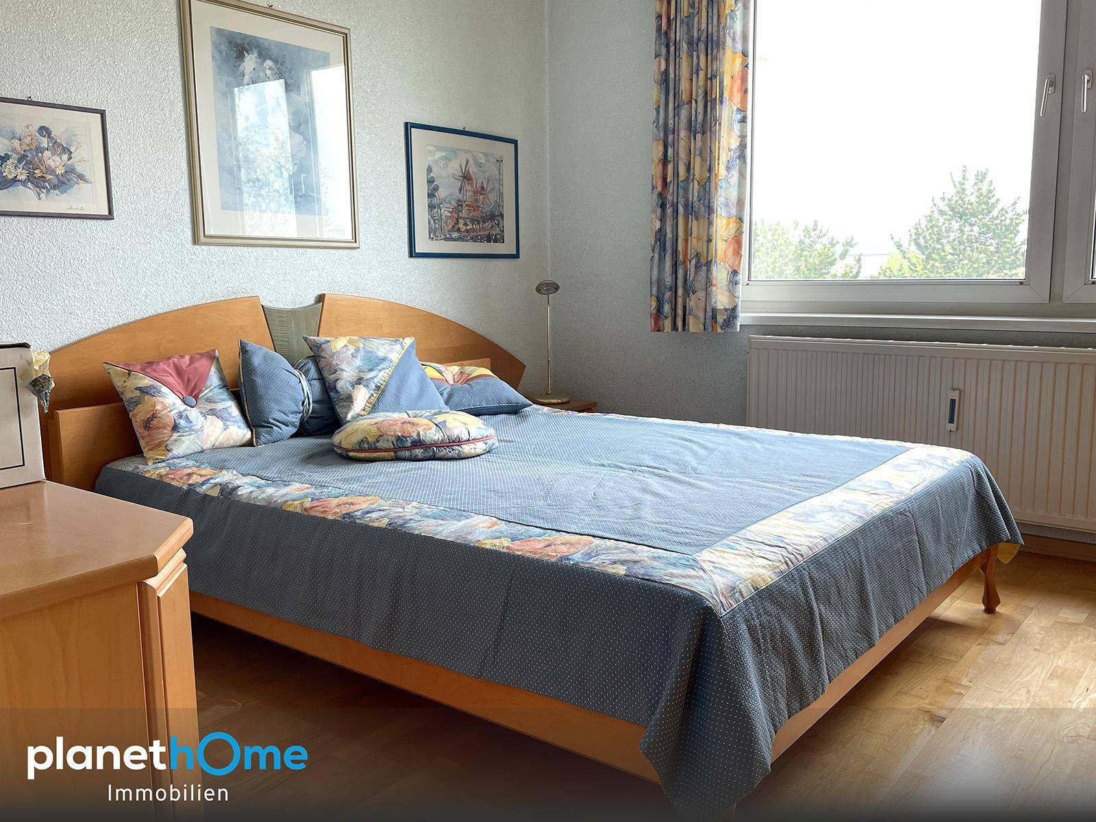 Helles Schlafzimmer mit einem Holzbett, blauer Bettwäsche und einem Fenster mit Blick ins Grüne.
