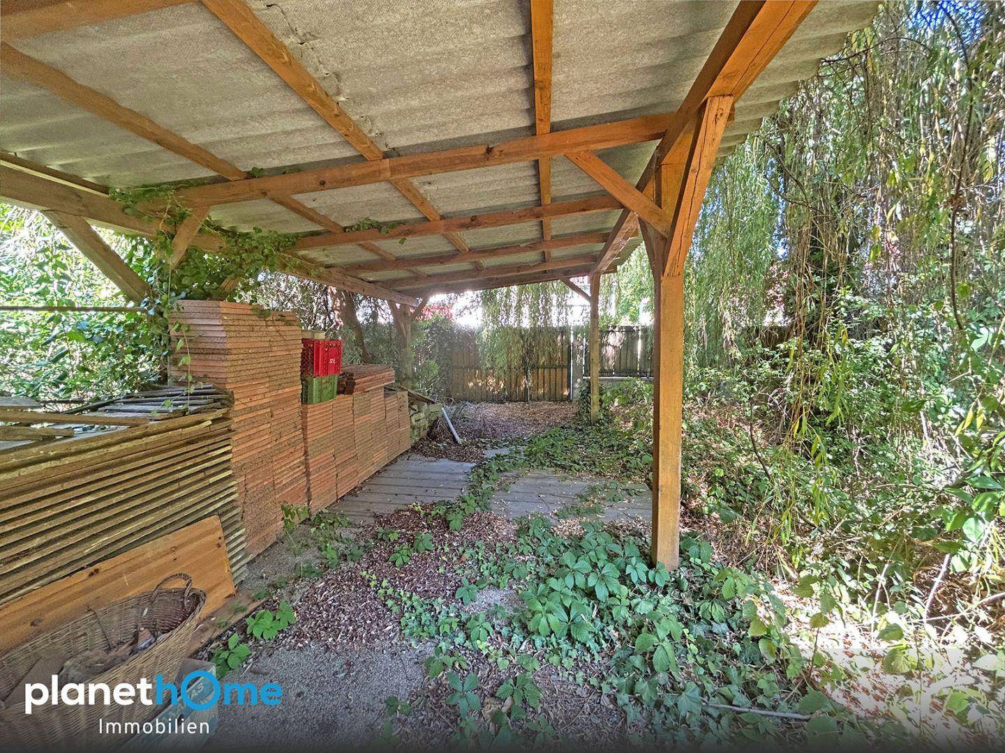 Holzunterstand im Gartenbereich, umgeben von üppigem Grün und gelagerten Materialien.