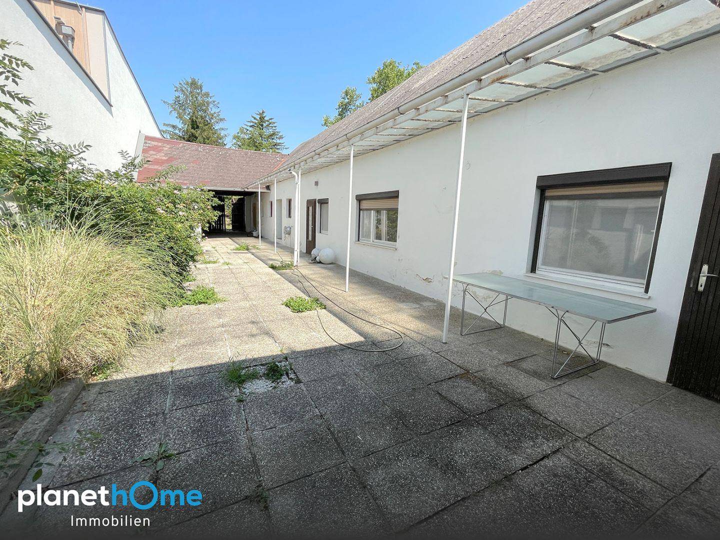 Lange gepflasterte Terrasse entlang des Gebäudes mit viel Grün und einem Tisch.