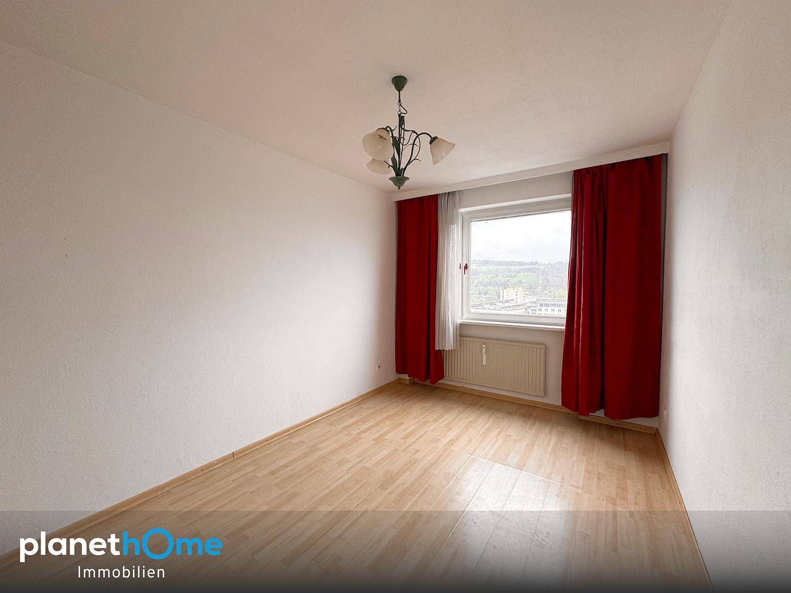 Leeres Zimmer mit Laminatboden, weißen Wänden und roten Vorhängen am Fenster.
