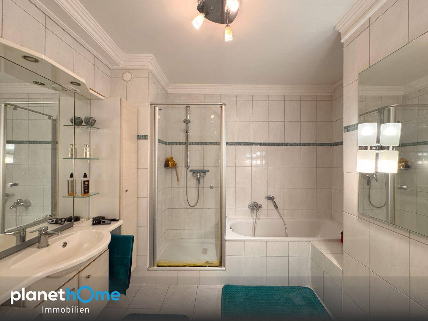 Badezimmer im Originalzustand mit weißen Fliesen, Badewanne mit Duschfunktion und Einzelwaschbecken.