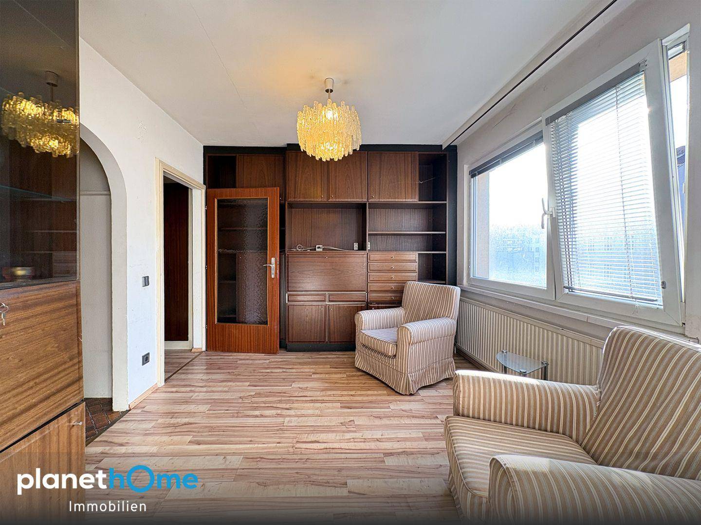 Heller Wohnraum mit Holzschrank, gemütlichem Sessel und Zugang zu einem weiteren Zimmer durch einen Bogen.