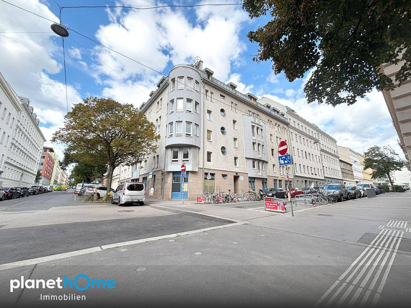 Eckgebäude in urbaner Umgebung mit geparkten Autos und Bäumen an der Straße.