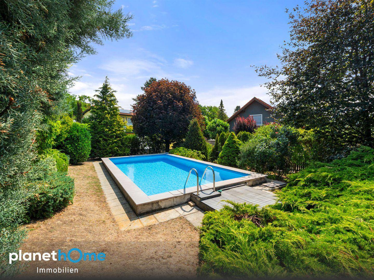 Rechteckiger Swimmingpool im Garten, umgeben von üppigem Grün und Bäumen unter blauem Himmel.