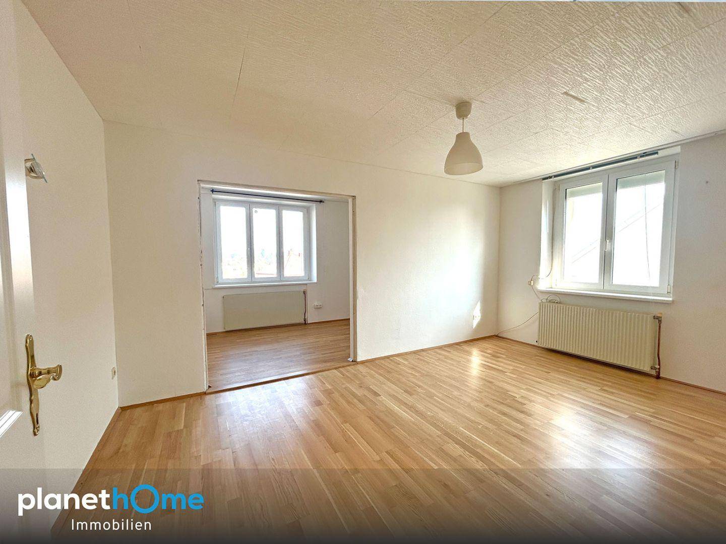 Helles, leeres Wohnzimmer mit Parkettboden und Zugang zu einem weiteren Raum.