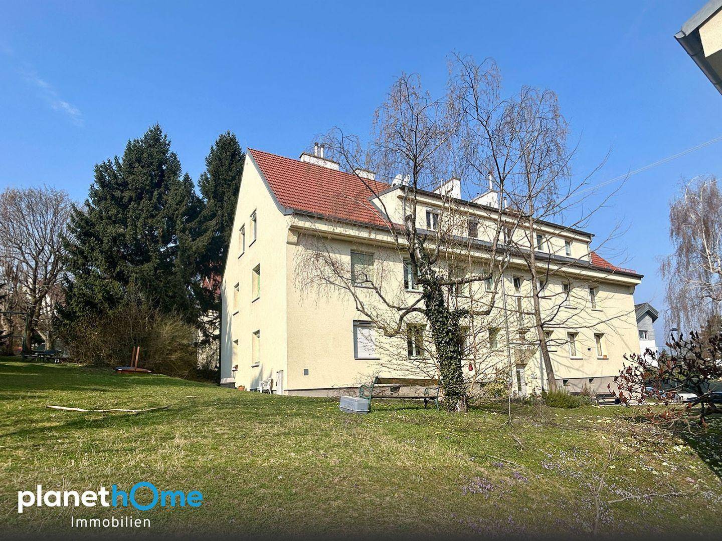 Mehrstöckiges Gebäude mit rotem Ziegeldach und großem Gartenbereich unter blauem Himmel.