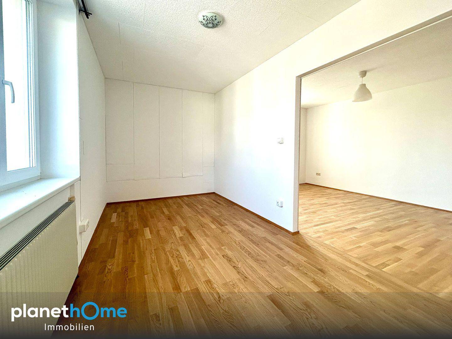 Leeres Schlafzimmer mit Parkettboden und Fenster, das zu einem weiteren Raum führt.