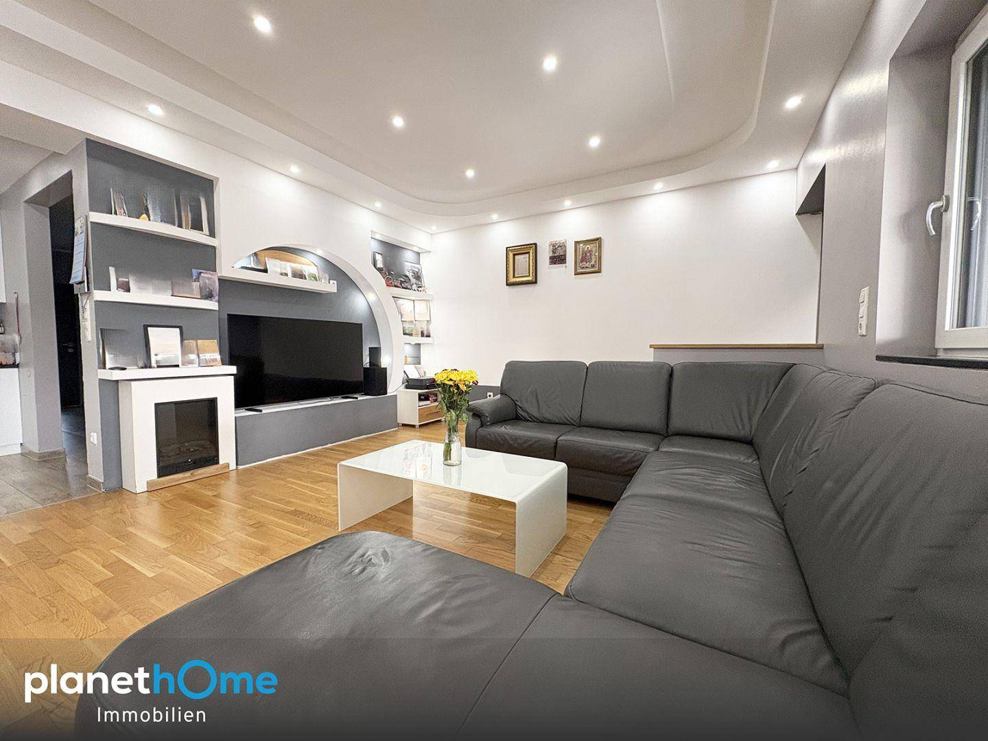Gemütliches Wohnzimmer mit großem Ecksofa, Holzboden und integrierten Regalen sowie Deckenstrahlern.