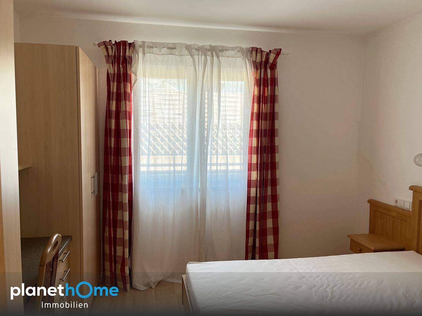 Schlafzimmer mit Bett, Kleiderschrank und Fenster mit rot-weiß karierten Vorhängen.