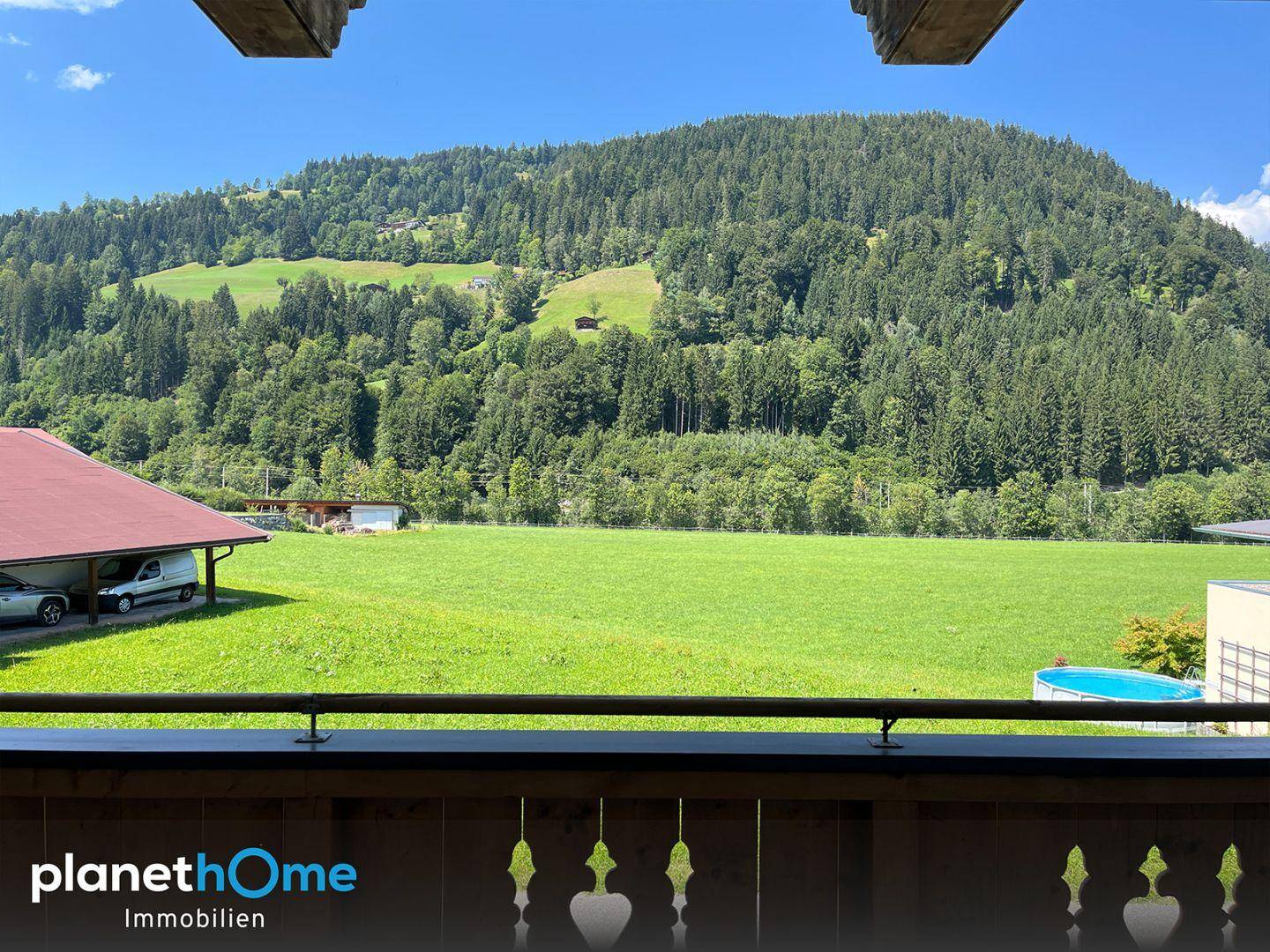 Blick vom Balkon auf weite grüne Wiesen, bewaldete Berge und einen kleinen Pool unter blauem Himmel.