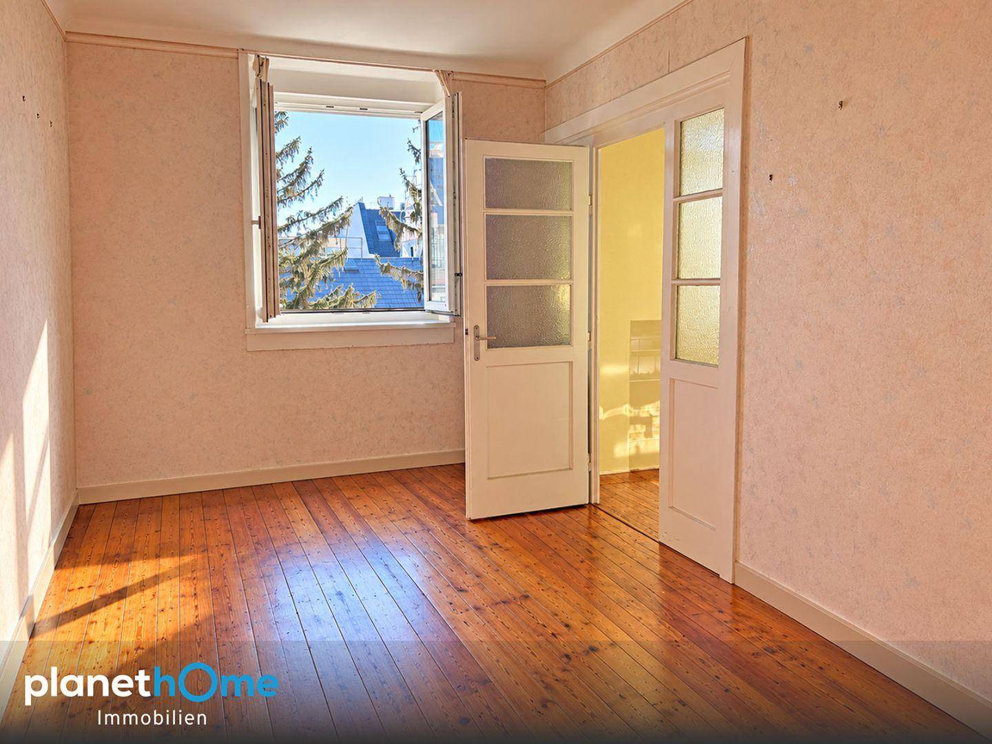 Freundliches Zimmer mit Holzboden, Fensterblick ins Grüne und Zugang zu einem weiteren Raum.