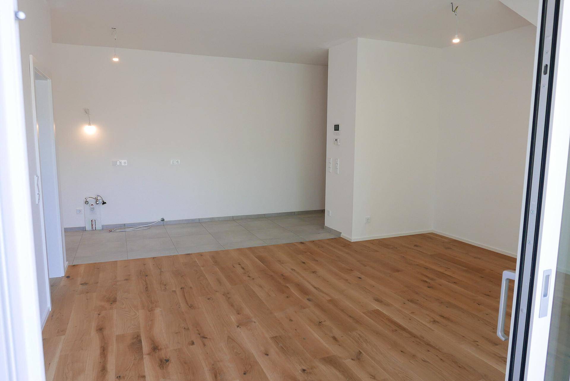 Offener Wohnbereich mit Holzboden und einem gefliesten Bereich, bereit für die Kücheninstallation.