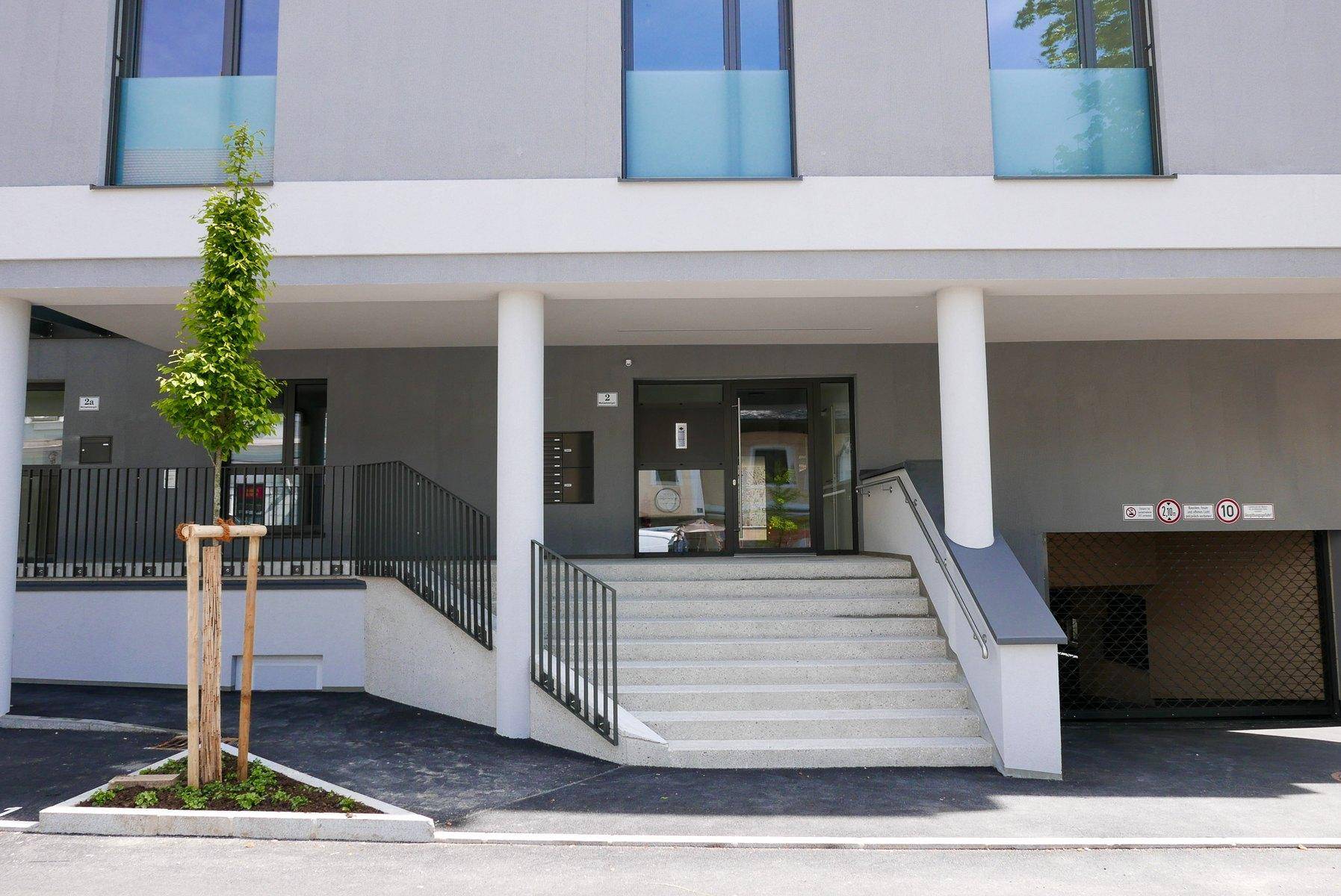 Eingangsbereich des modernen Gebäudes mit Treppe, Rampe und Tiefgaragenzufahrt, barrierefrei gestaltet.