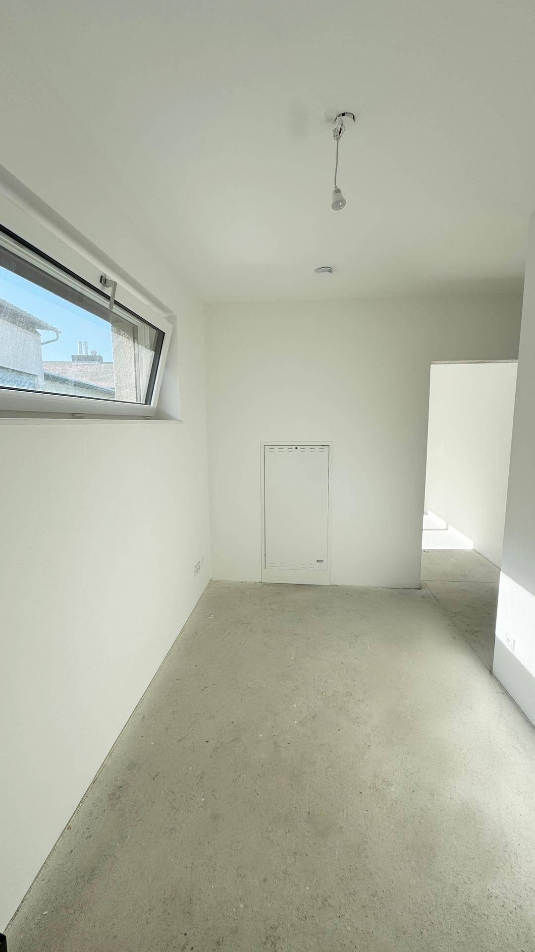 Unfertiger Raum mit weißen Wänden, Betonboden und einem kleinen Fenster, bereit für den Ausbau.