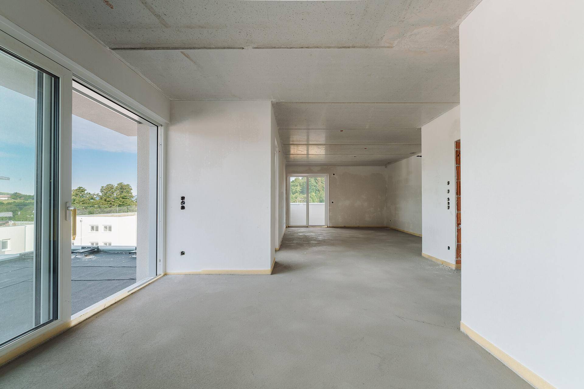 Geräumiger, unfertiger Innenraum mit großen Fenstern und Zugang zu einem Außenbereich.