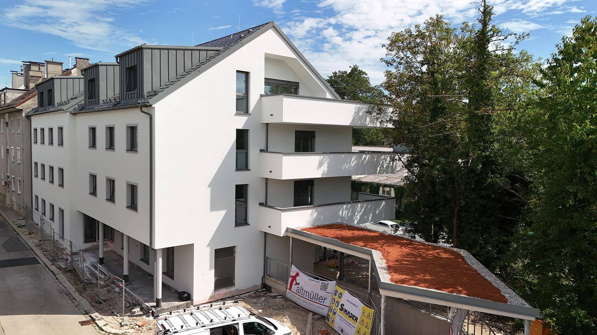 Seitenansicht des Neubaus mit weißer Fassade, Balkonen und begrüntem Dachbereich.
