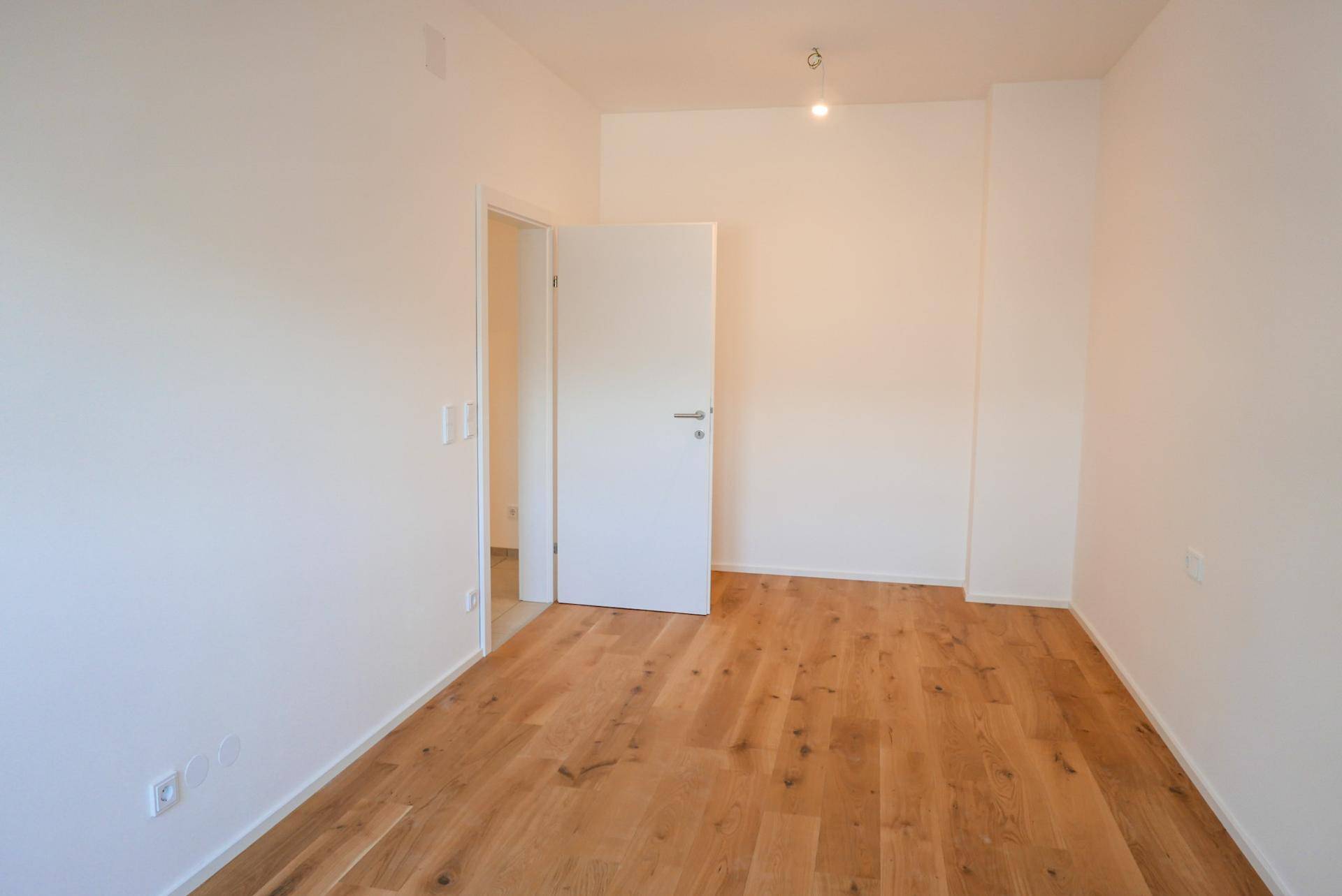 Helles, leeres Zimmer mit Holzboden und weiß gestrichenen Wänden.