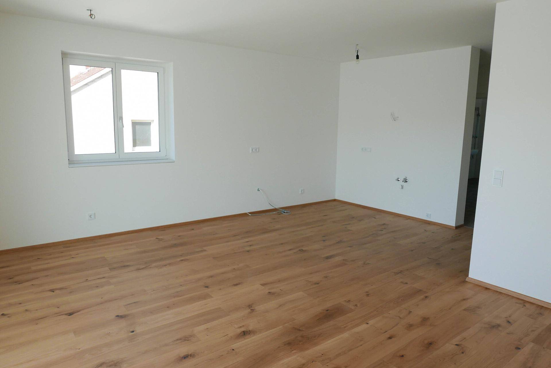 Leeres Wohnzimmer mit Holzboden, weißen Wänden und großem Fenster.