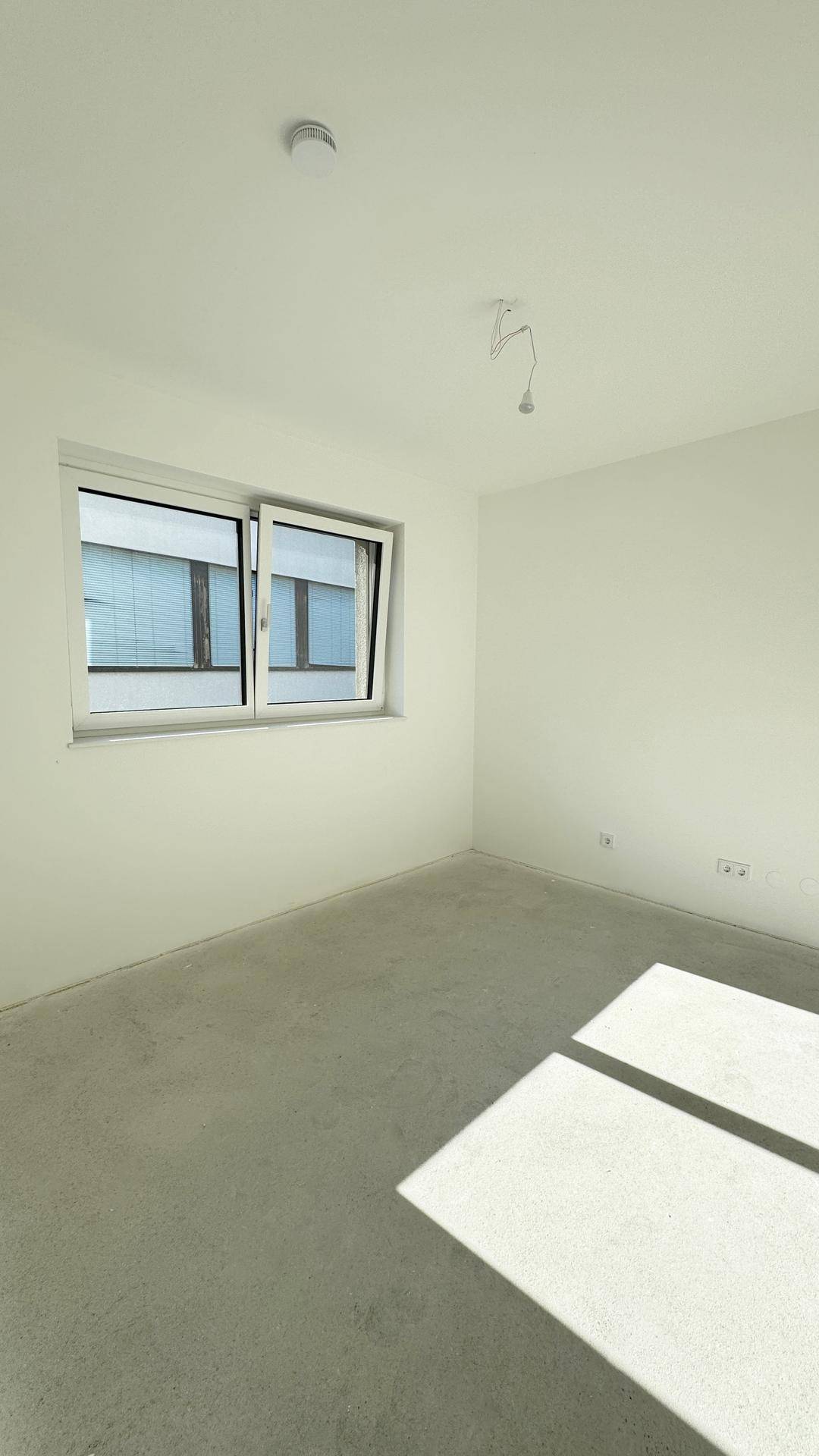 Leerer Raum im Rohbau mit weißen Wänden, Betonboden und einem Fenster, das Tageslicht hereinlässt.
