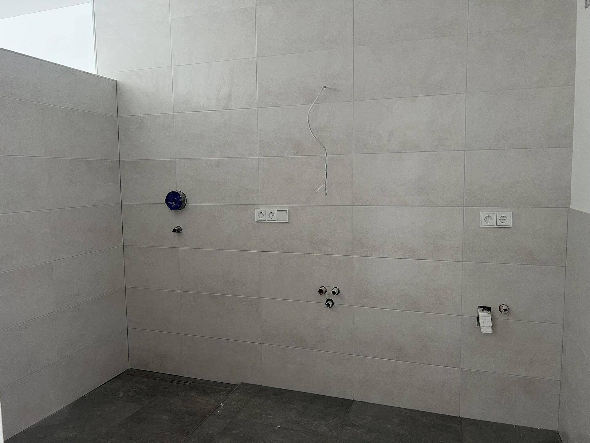 Badezimmer im Rohbau mit hellgrauen Wandfliesen und sichtbaren Anschlüssen für Sanitäranlagen.