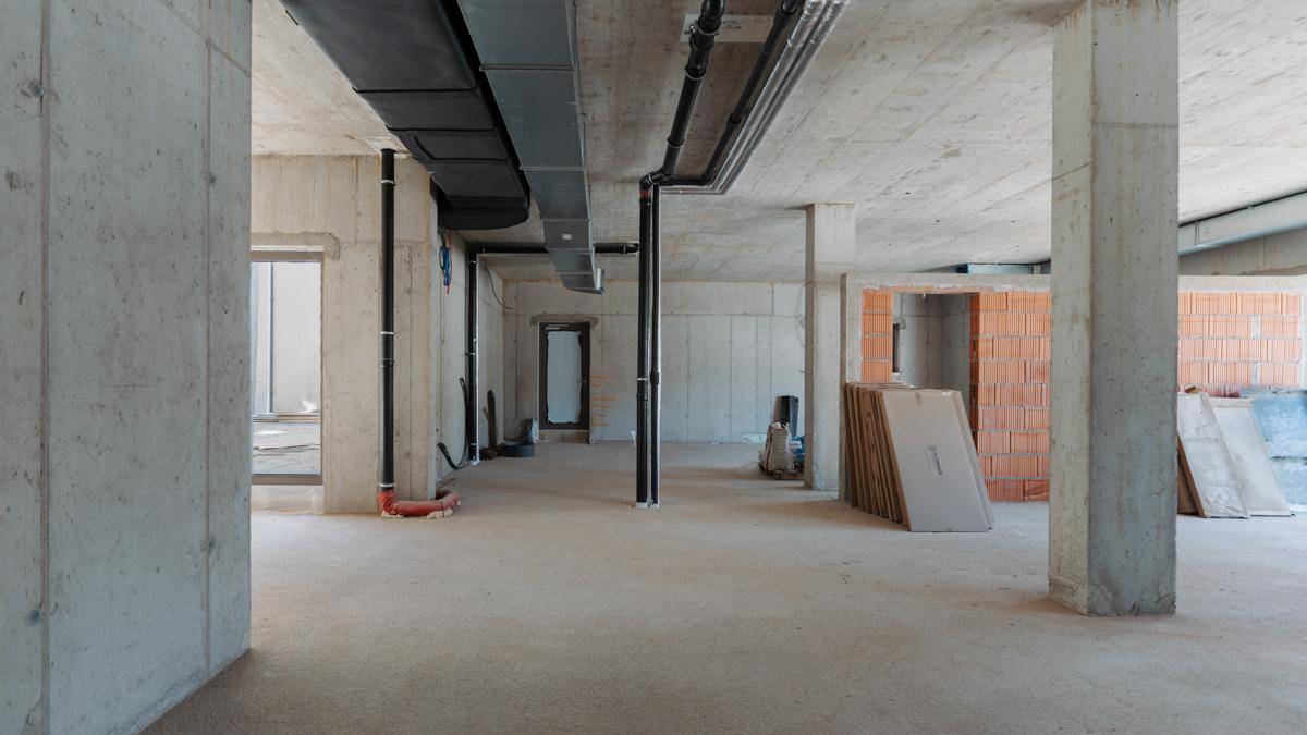 Großer, unfertiger Kellerbereich mit Betonstützen und -wänden, sichtbaren Installationen und Baumaterialien.