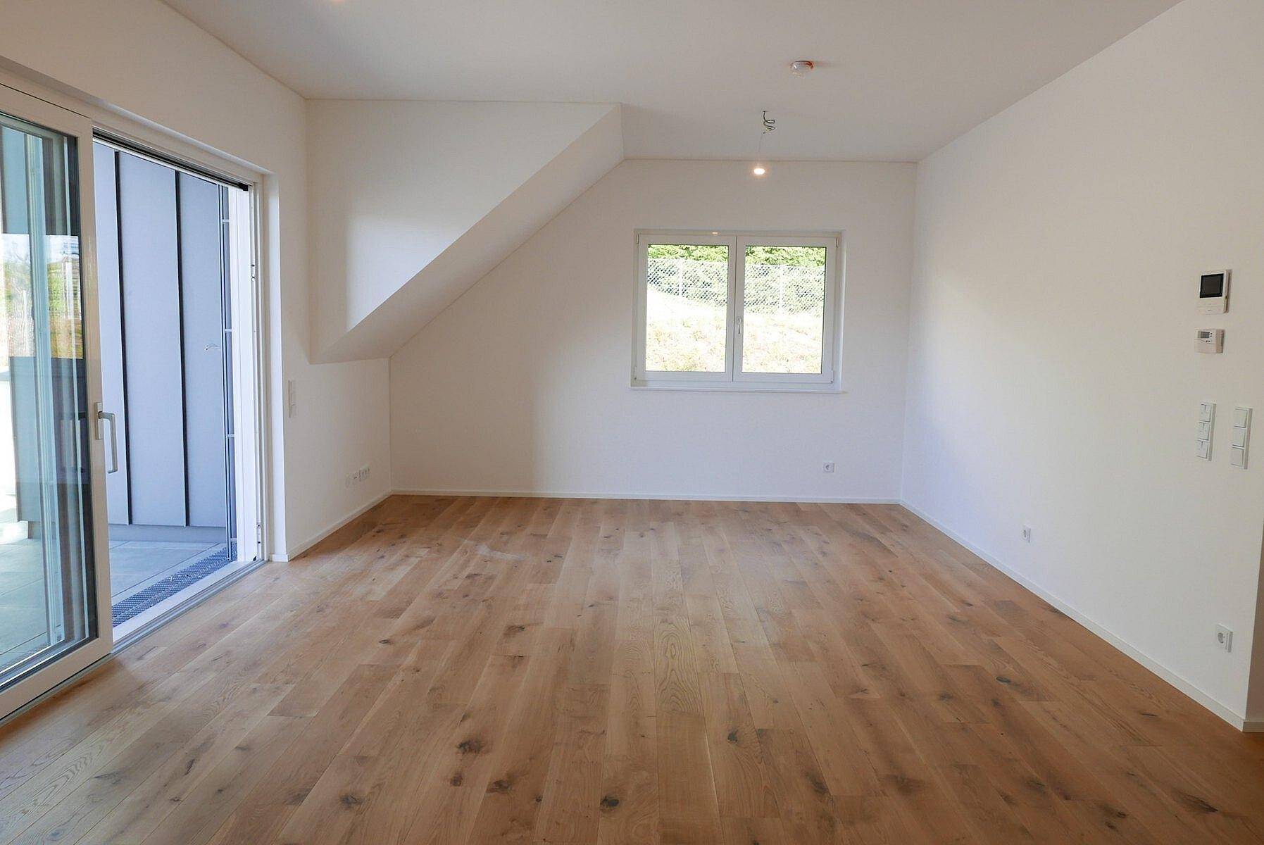 Geräumiger Raum mit Holzboden, großen Fenstern und Zugang zu einem Balkon oder einer Terrasse.
