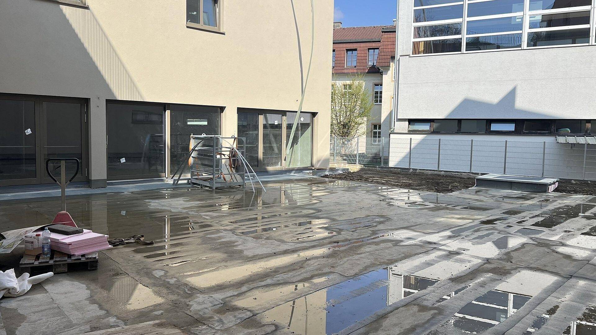 Außenbereich eines Neubaus mit großer, nasser Terrassenfläche und angrenzenden Gebäuden.