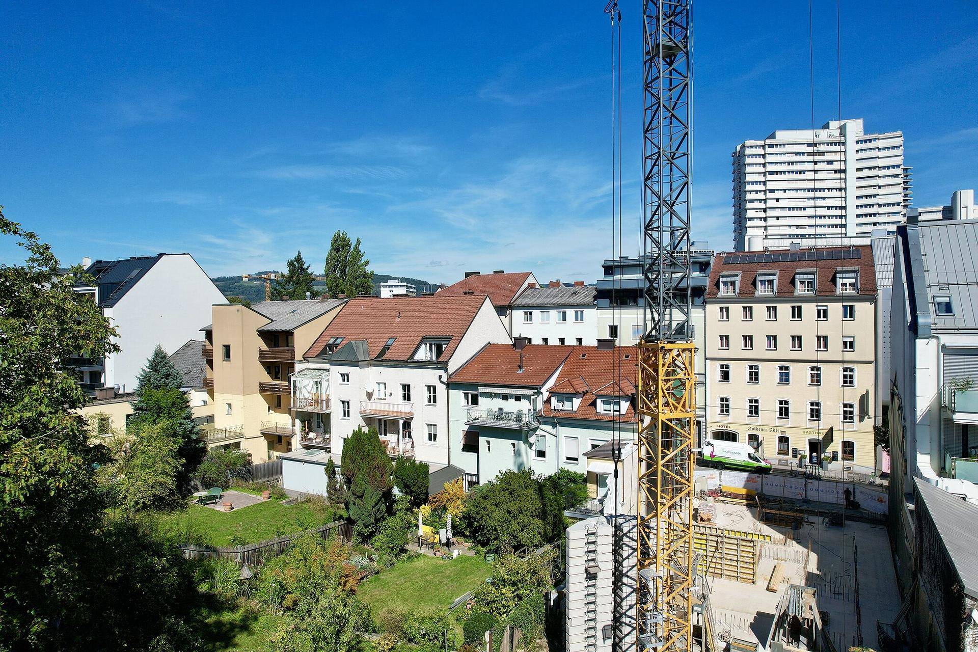Baustelle mit Kran und bestehenden Gebäuden, darunter ein Hochhaus, unter klarem blauem Himmel.