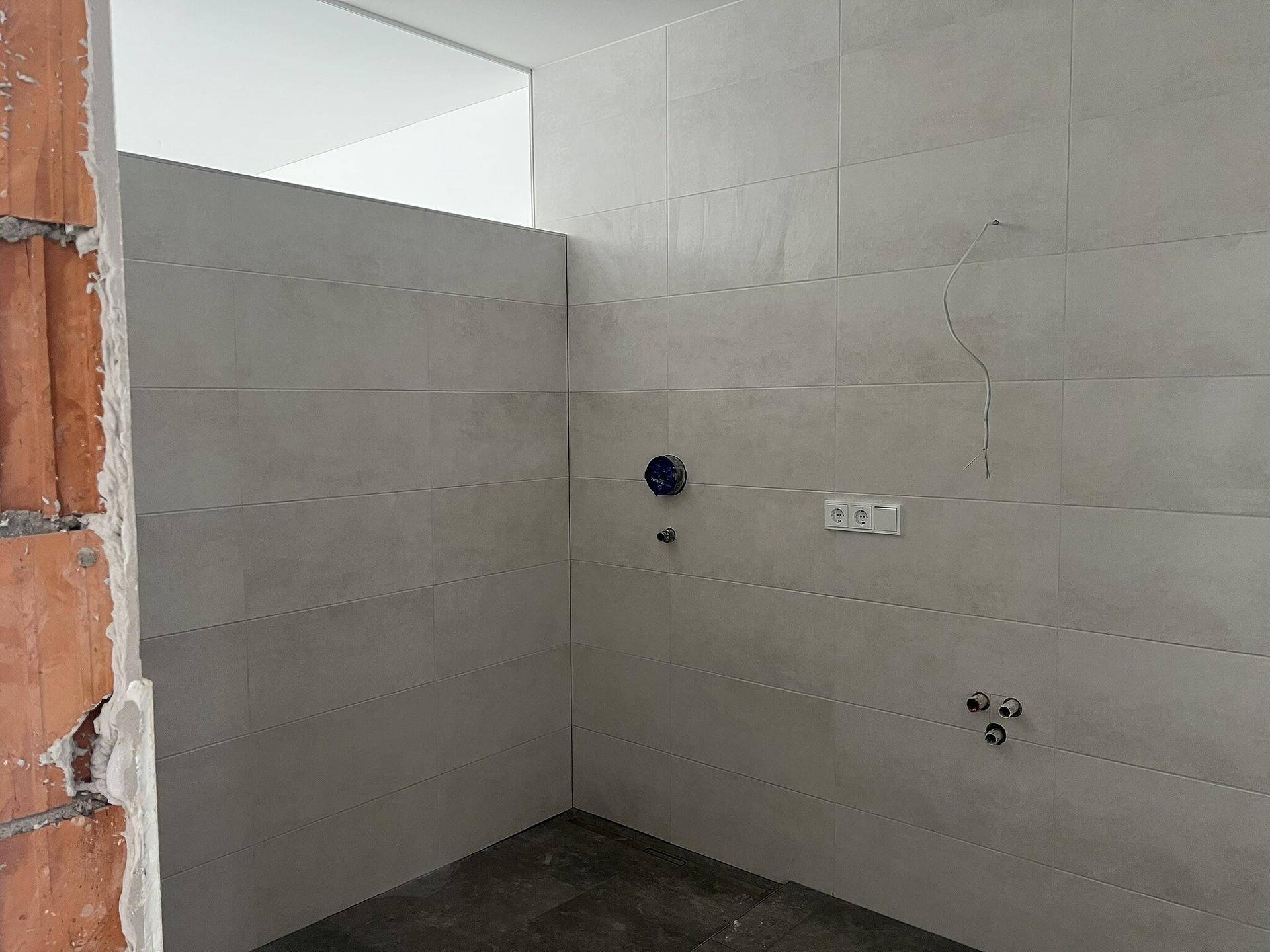 Badezimmer im Rohbau mit hellgrauen Wandfliesen und sichtbaren Anschlüssen für Sanitäranlagen.