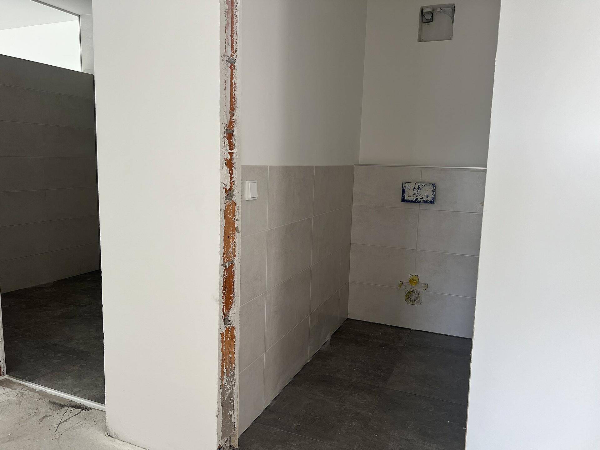 Badezimmer im Rohbau mit grauen Fliesen an den Wänden und dunklem Boden.