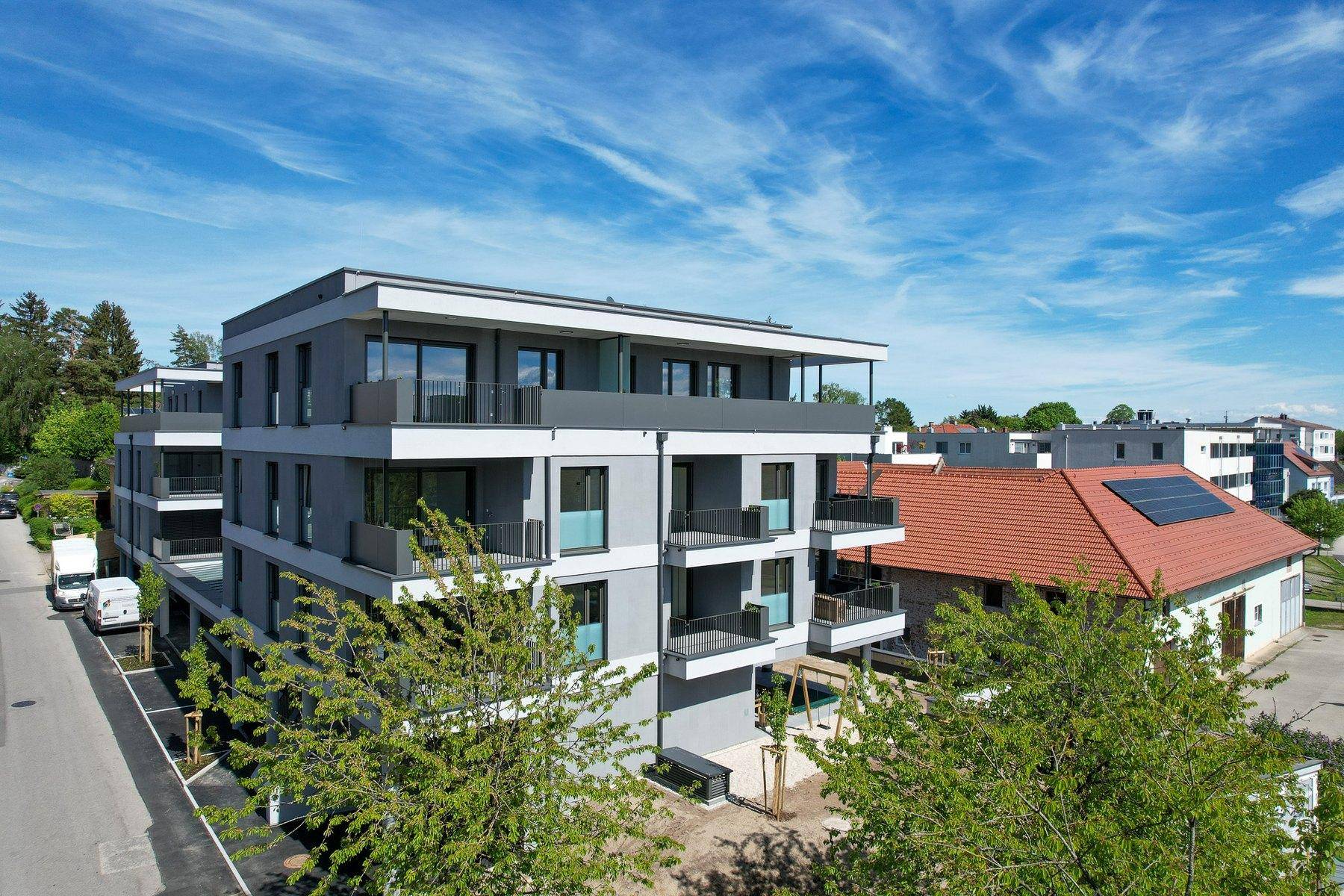 Modernes Mehrfamilienhaus mit Balkonen und Photovoltaikanlage auf dem Dach, in grüner Umgebung.