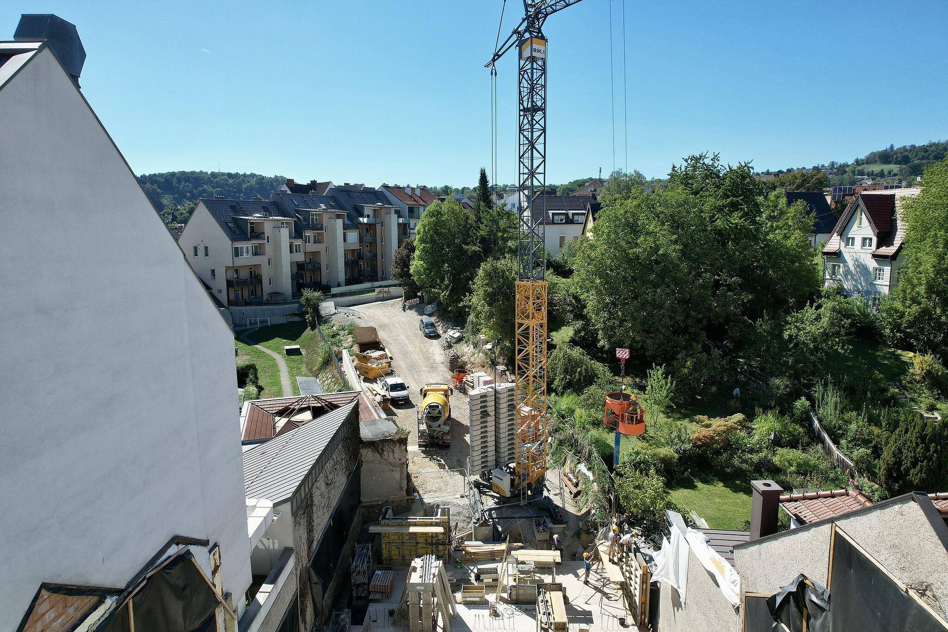 Baustelle mit Kran und umliegender Wohnbebauung in einer grünen Umgebung unter blauem Himmel.