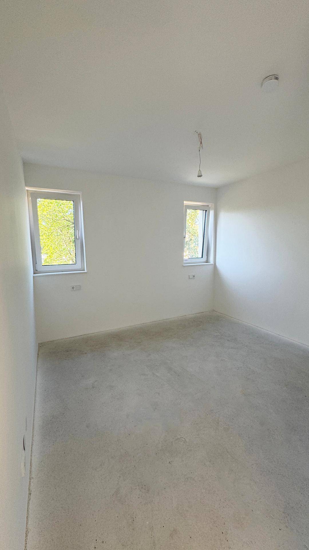 Leerer Raum im Rohbau mit weißen Wänden, Betonboden und zwei Fenstern für ausreichend Helligkeit.