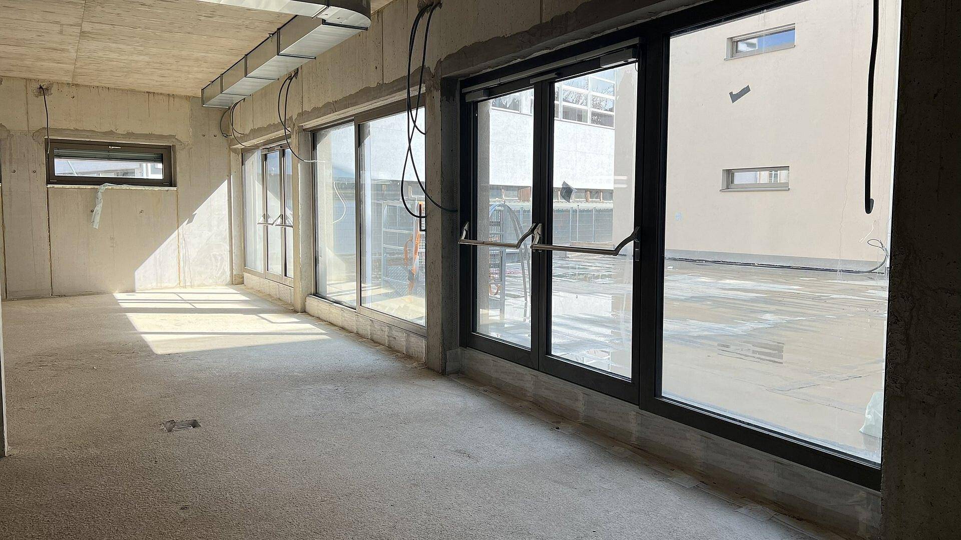 Heller Rohbau-Innenraum mit großen Fensterfronten und sichtbaren Installationen, die viel Tageslicht hereinlassen.