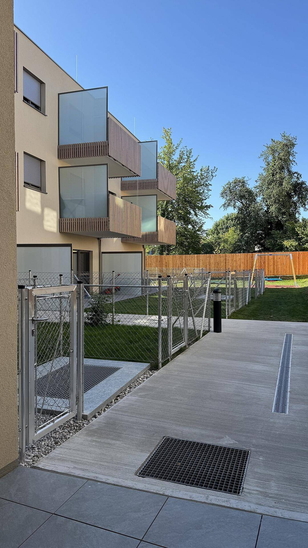 Moderner Gebäudekomplex mit Balkonen und einem eingezäunten Gartenbereich mit Spielplatz im Hintergrund.