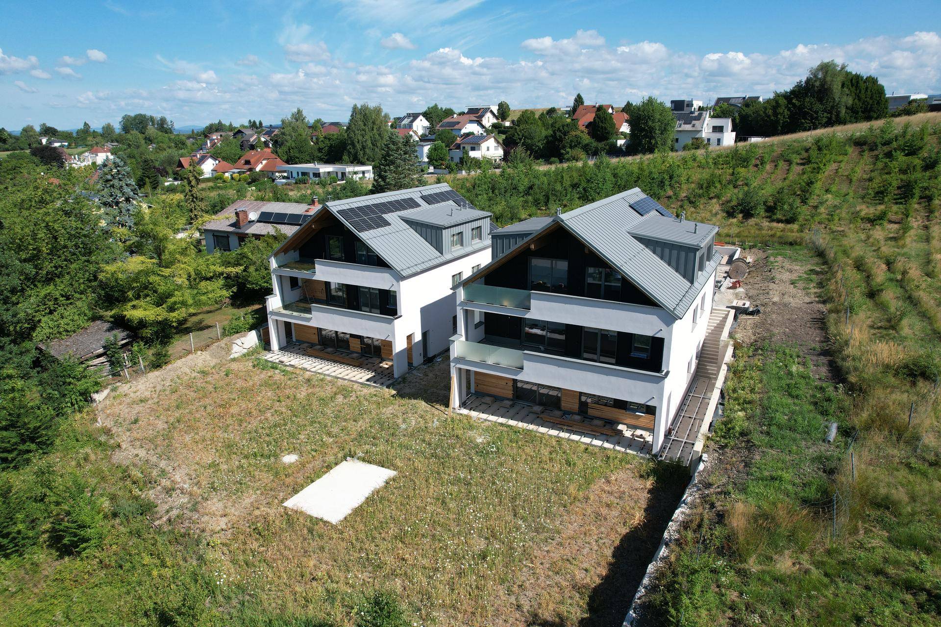 Zwei moderne Neubauhäuser mit Photovoltaikanlagen in einer grünen, hügeligen Landschaft.